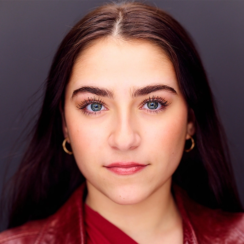 A young woman with long dark hair and striking blue eyes, reminiscent of Vita Augugliaro, stares directly at the camera. She has full eyebrows, light makeup, gold hoop earrings, and wears a red top with a matching red leather jacket.