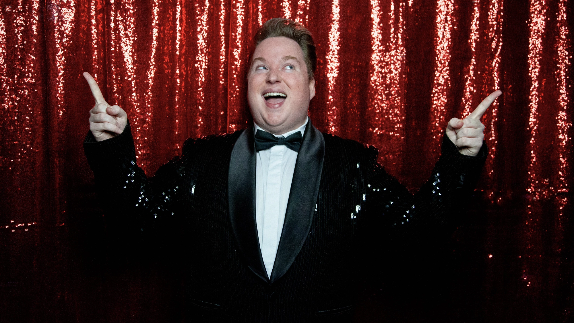Macon Prickett - Show Art A smiling person in a black sequin tuxedo and bow tie stands in front of a sparkling red sequin curtain, pointing upwards with both hands and looking slightly up, capturing the excitement and joy of The Comeback.