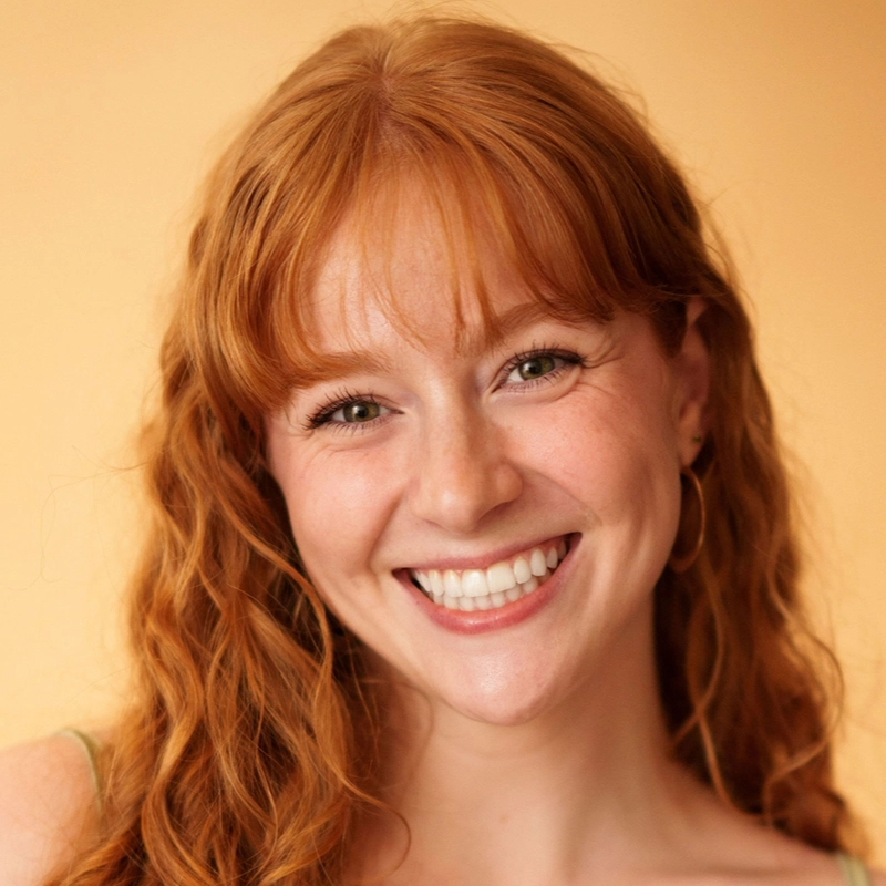 A young woman with long, wavy red hair and fair skin smiles warmly at the camera. She has bangs and is wearing small hoop earrings. The background is a soft, solid warm beige color, giving the image a bright and inviting feel.