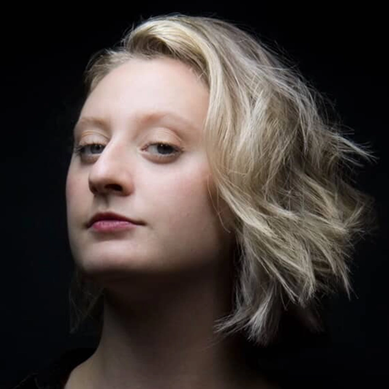 A person with short, wavy blonde hair looks slightly upward with a confident expression. They have fair skin and light-colored eyes. The background is black, making their face and hair stand out in dramatic lighting from the left.
