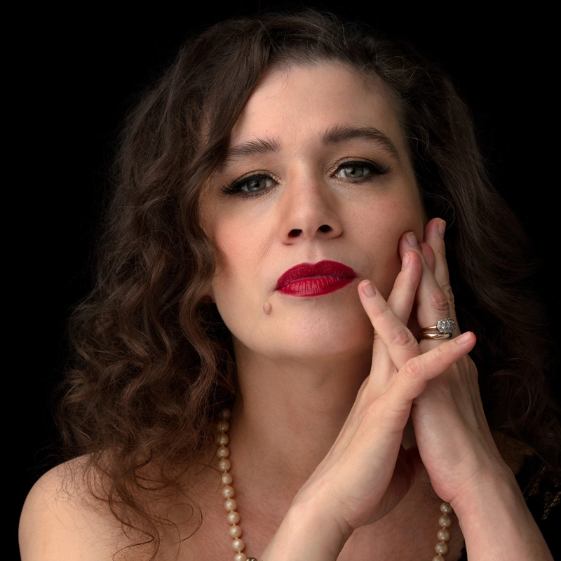 A woman with wavy brown hair, bright red lipstick, and a beauty mark on her cheek gazes forward. Reminiscent of Maude Maggart, she wears a pearl necklace, an engagement ring, and rests her chin and cheek on her hands against a black background.