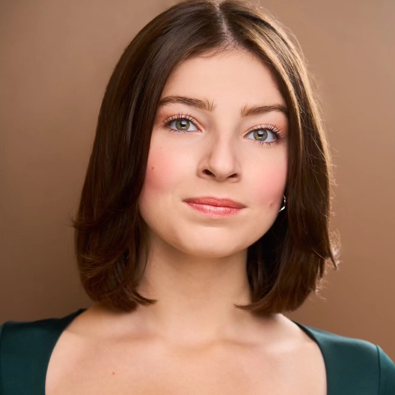 A young woman with fair skin and straight, shoulder-length brown hair poses against a brown background. She has Majella-green eyes, subtle pink makeup, and wears a dark green top with a wide neckline, looking confidently and slightly smiling at the camera.