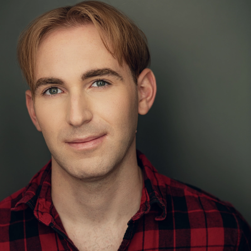 A young adult with light skin, blue eyes, and short strawberry-blond hair, parted in the middle, smiles softly. Mikey Marmann wears a red and black plaid shirt and poses against a plain, dark gray background. The lighting is warm and even.