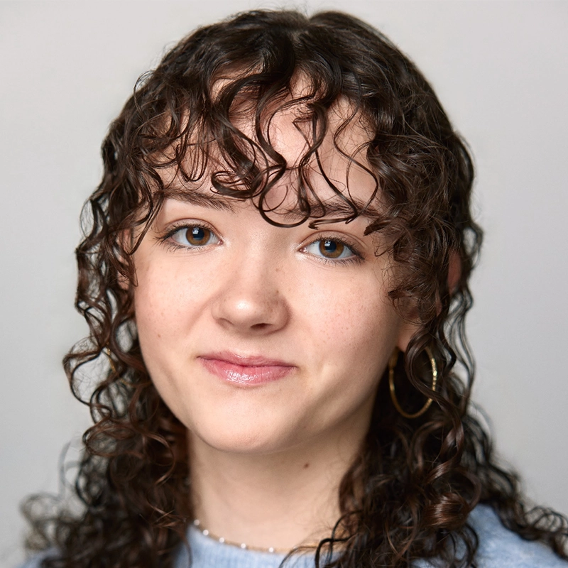 Vita Augugliaro, a young person with light skin and blue eyes, smiles gently at the camera. They have brown, shoulder-length curly hair with bangs and wear a light blue top, pearl necklace, and gold hoop earring against a plain background.