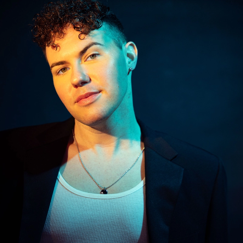 Brendan McCann, with short curly hair and earrings, wears a black blazer over a white tank top and a silver necklace. Teal and yellow lighting highlights his face and shoulders against a dark background as he looks calmly into the camera.