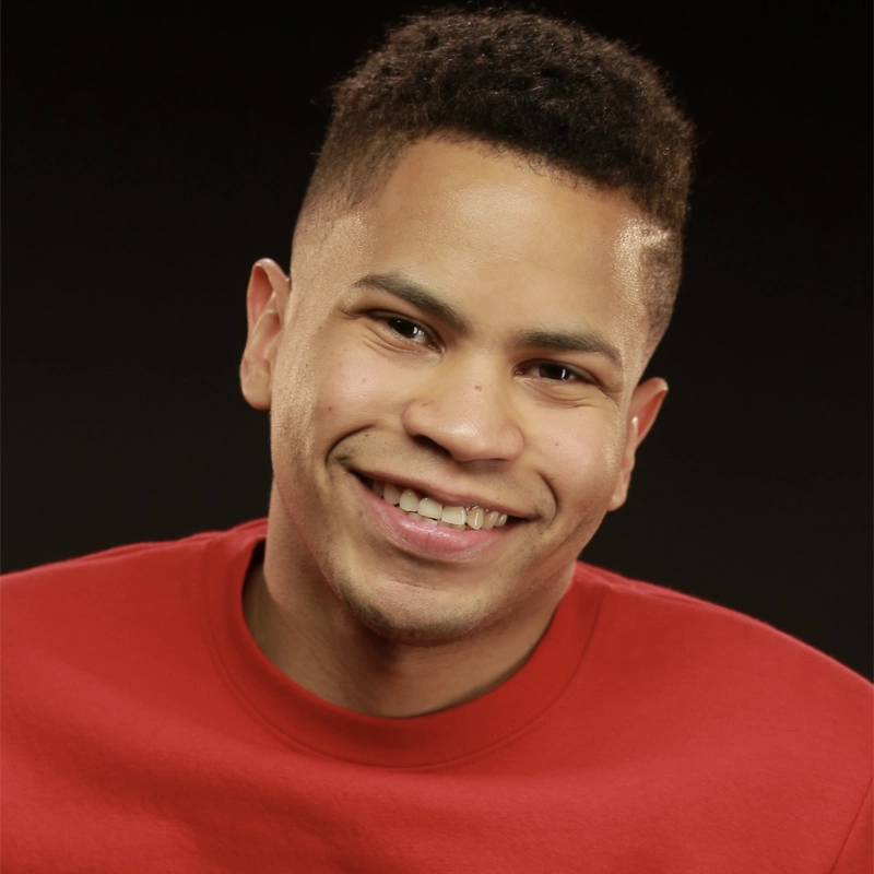 Sylvester McCracken III, a young man with short curly hair, smiles warmly at the camera. He wears a bright red crew-neck shirt and sits against a plain black background, the lighting highlighting his friendly expression and smooth complexion.