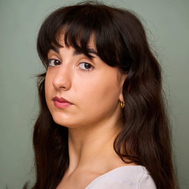 A young woman with long wavy brown hair and bangs, Allison Mele, looks into the camera with a neutral expression. She is wearing a white top, small gold hoop earrings, and has light, natural makeup against a plain muted green background.