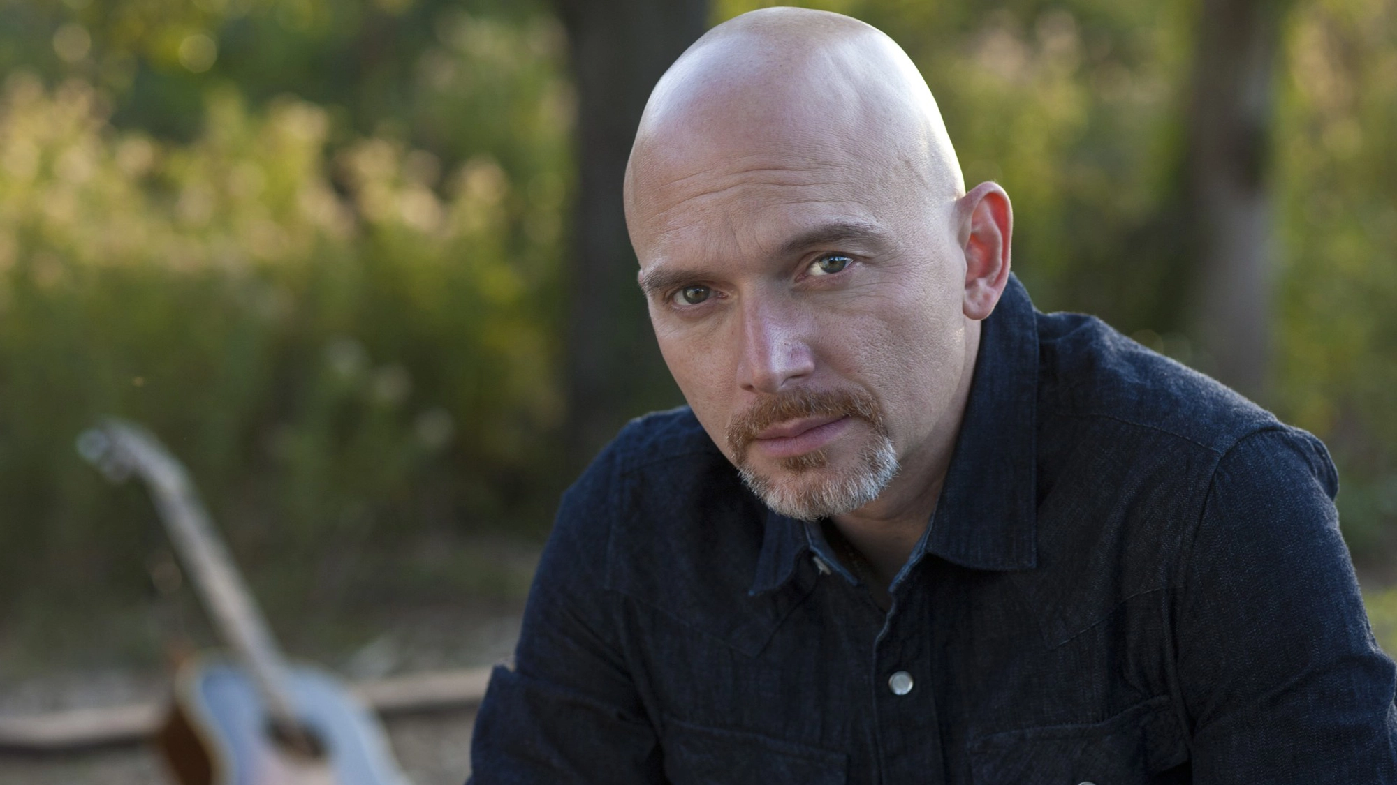A bald man with a trimmed goatee and mustache wears a dark denim shirt, gazing seriously at the camera. Songsmith Michael Cerveris stands outdoors with blurred greenery and a guitar in the background, suggesting a natural, relaxed setting.