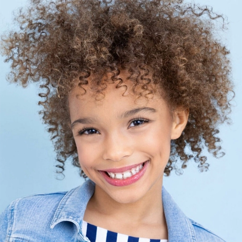 A smiling child with curly brown hair wears a denim jacket over a navy and white striped top. The background is light blue, and the child’s wide smile shows missing front teeth. The overall mood is cheerful and vibrant.