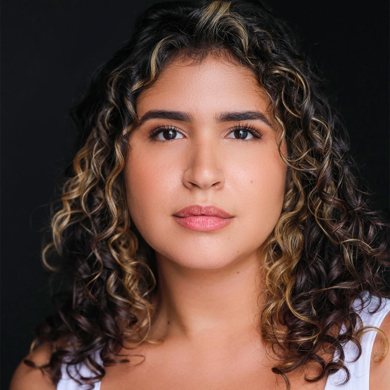 Annabell Mizrahi, a young woman with medium skin and curly, shoulder-length hair featuring blonde highlights, poses against a dark background. She wears a white sleeveless top and looks directly at the camera with a neutral, confident expression.