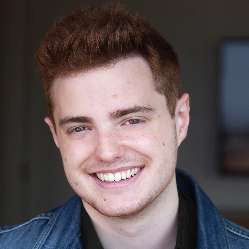 A young man with short, light brown hair and fair skin is smiling warmly. He is wearing a denim jacket over a dark shirt. The background is softly blurred, with neutral colors and a rectangular object on the wall behind him.