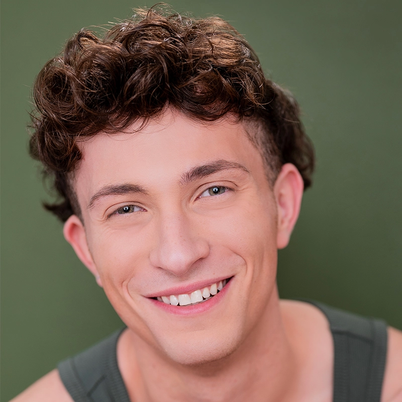 A young man with short, curly brown hair and blue eyes smiles warmly at the camera. He wears a dark green tank top and is posed in front of a plain olive green background. The lighting is soft, highlighting his clear skin and cheerful expression.