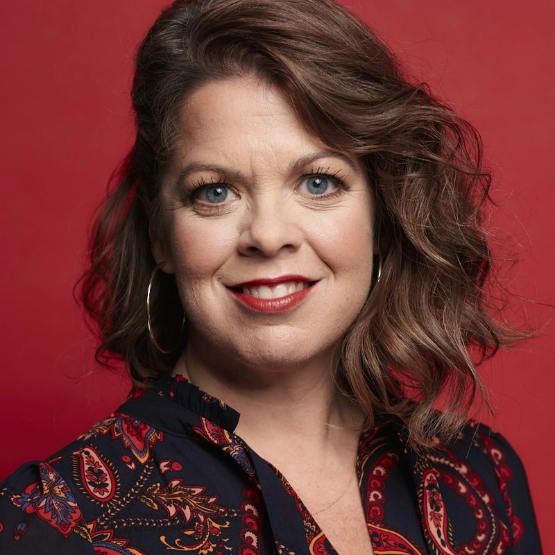 Christine O’Grady, with wavy brown hair and blue eyes, smiles at the camera. She wears large hoop earrings, red lipstick, and a dark blouse with a colorful paisley pattern against a solid red background.