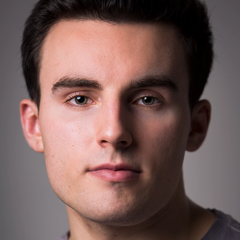 Connor O’Shea, a young man with short dark hair and fair skin, looks directly at the camera with a neutral expression. He has light-colored eyes, well-groomed eyebrows, and slight stubble. The softly lit background focuses attention on his face.