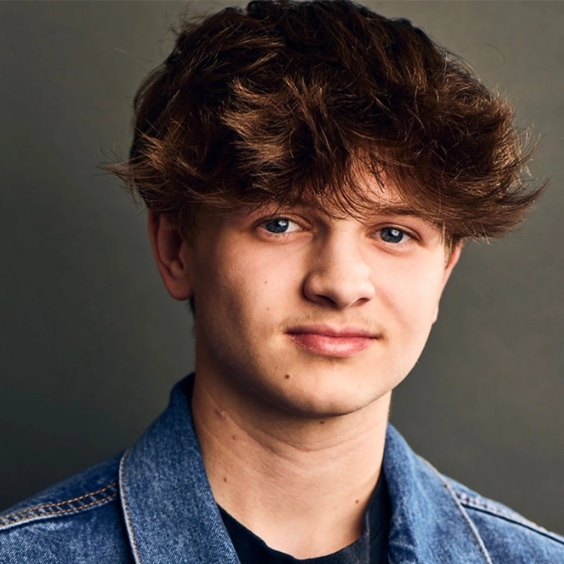 A teenage boy with wavy brown hair and blue eyes looks at the camera with a slight smile. He wears a blue denim jacket over a dark shirt and stands against a plain, dark background. The lighting highlights his facial features.