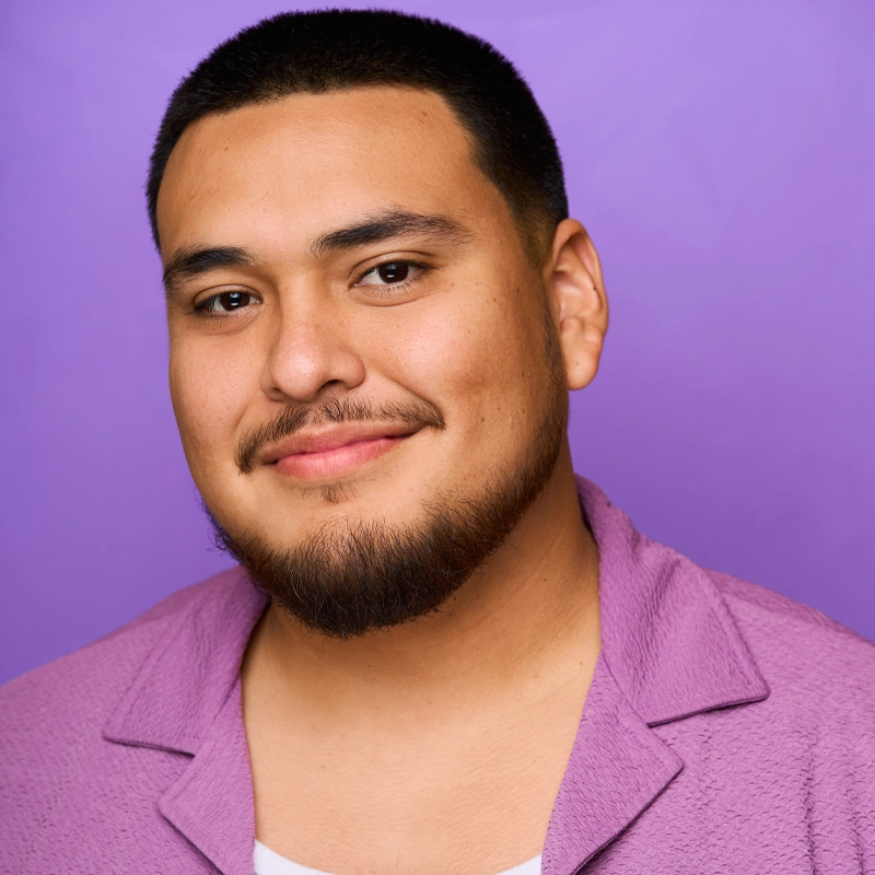Carlos Peréz, a man with short black hair and a trimmed beard, smiles gently at the camera. He is wearing a textured purple shirt over a white top, set against a solid lavender background for a coordinated and friendly portrait.