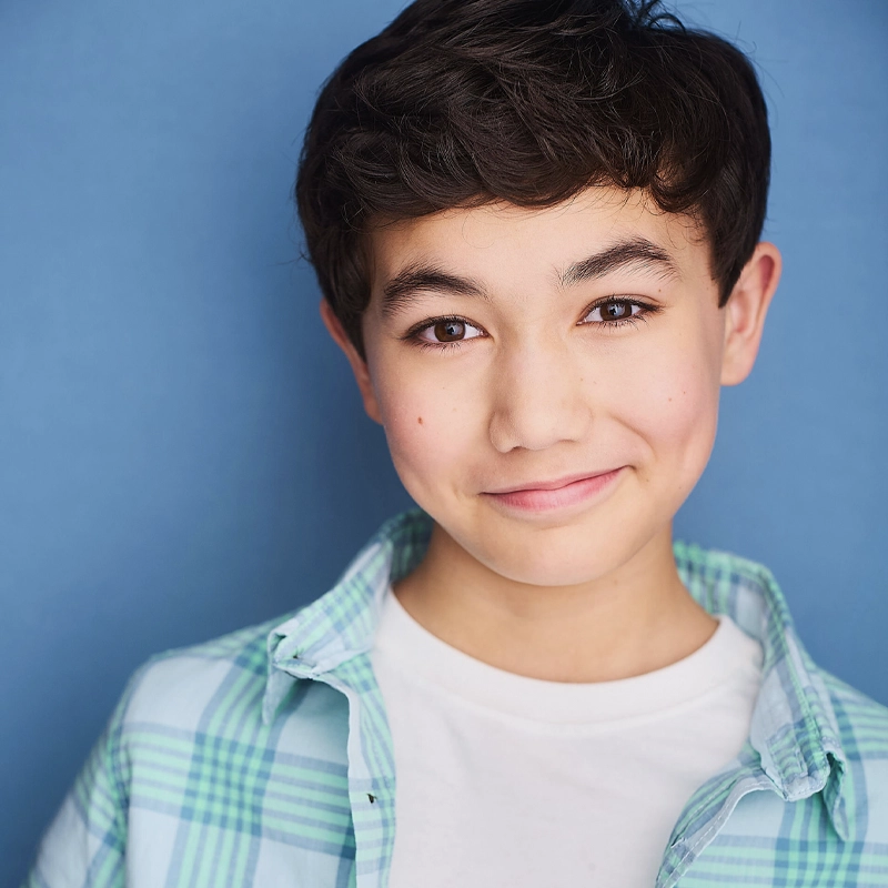 A smiling young boy with short dark hair poses against a blue background. He wears a teal and white plaid shirt over a plain white T-shirt. He looks directly at the camera with a friendly expression.