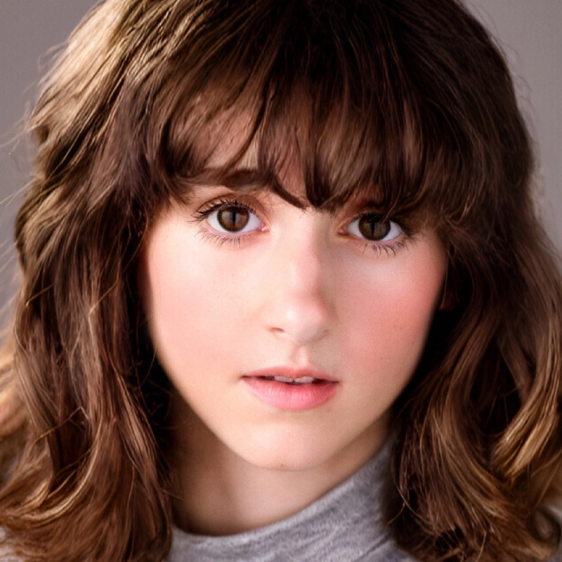 A person with wavy brown hair and bangs looks directly at the camera. They have fair skin, large brown eyes, and natural makeup with rosy cheeks and pink lips. The softly blurred background gives an Iona Press portrait feel as they wear a light gray top.