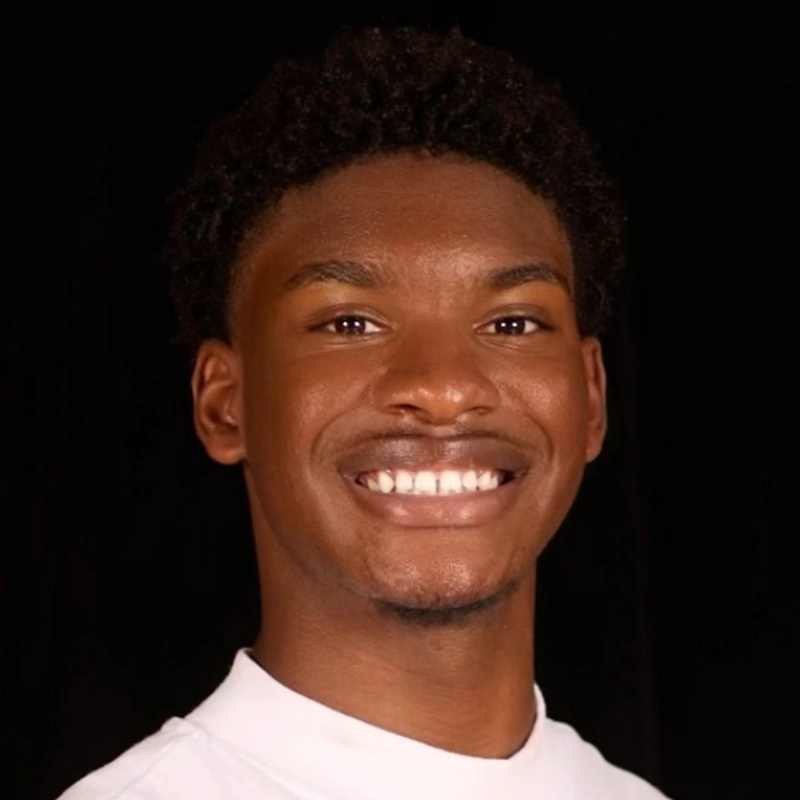 A young man with short curly hair smiles warmly at the camera. He is wearing a white shirt and is posed against a solid black background. The lighting highlights his face, giving the image a friendly and inviting feel.