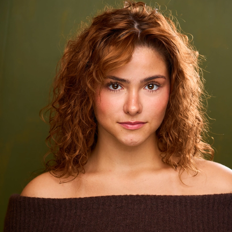 Riley Quinn-Caruso, a young woman with curly, reddish-brown hair and light skin, gazes directly at the camera in a brown off-the-shoulder sweater. Soft lighting and an olive green background create a warm, inviting atmosphere.