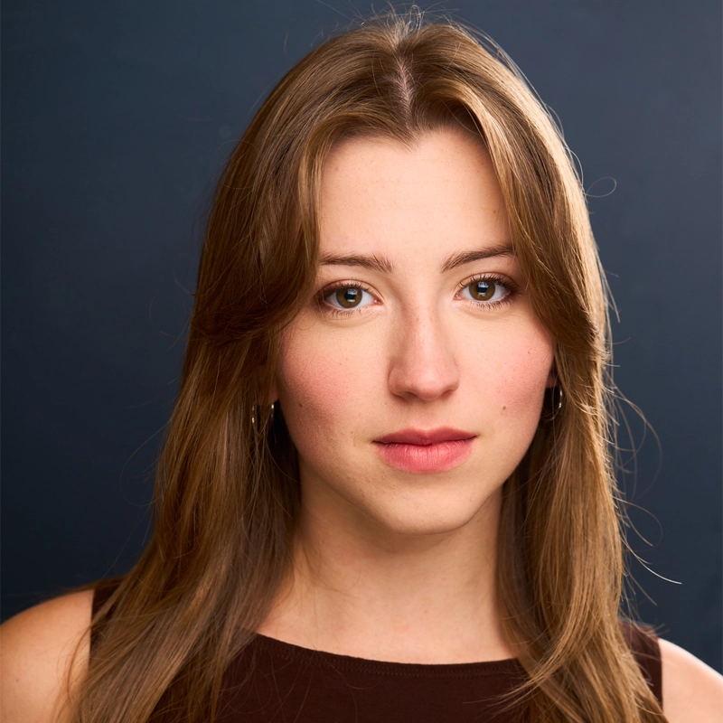 Jessica Ragucci, a young woman with straight, light brown hair and fair skin, looks directly at the camera. She wears a sleeveless dark top and subtle makeup against a plain, dark blue background, creating a professional headshot appearance.