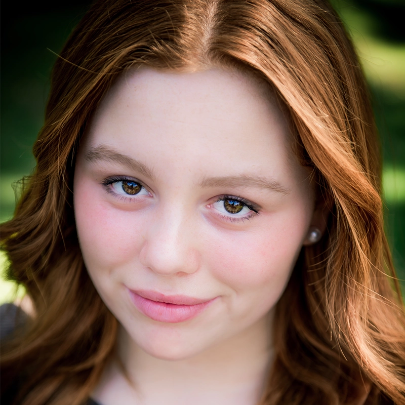 A young person with fair skin, long reddish-brown hair, and hazel eyes looks directly at the camera, smiling gently. The background is softly blurred green, suggesting an outdoor setting with natural light.