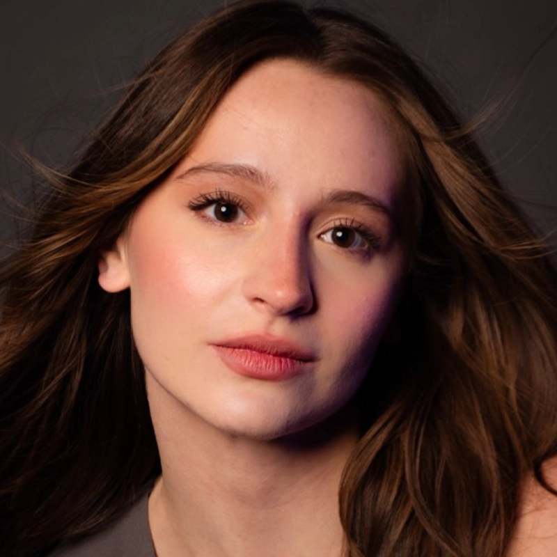 A young woman with long brown hair and fair skin, resembling Jordan Reising, looks into the camera with a neutral expression. She wears natural makeup, pink blush, and light lipstick. Her softly blowing hair frames her face against a dark background.