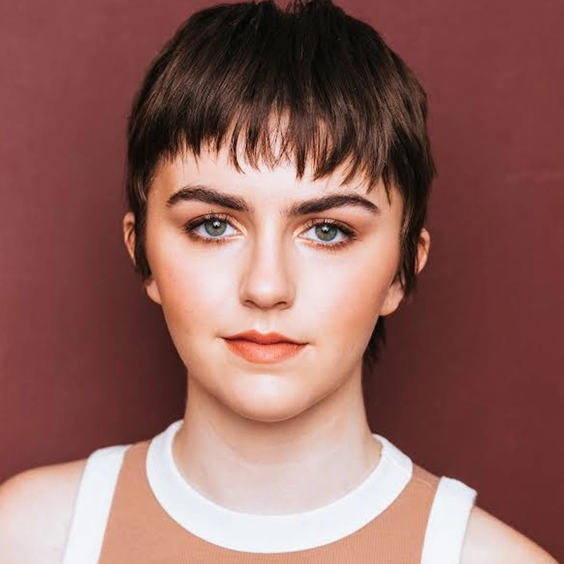 Corinne Renick, a young person with short, dark brown hair styled in a textured pixie cut, faces the camera. They have fair skin, bold eyebrows, light eye makeup, and peach lipstick, wearing a sleeveless beige and white top against a maroon background.