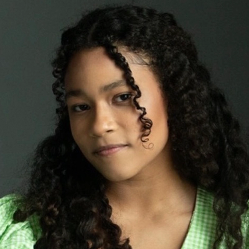A young person with medium brown skin and long, curly dark hair looks softly at the camera. They wear a light green, checked top and have a relaxed, gentle expression against a plain dark background.