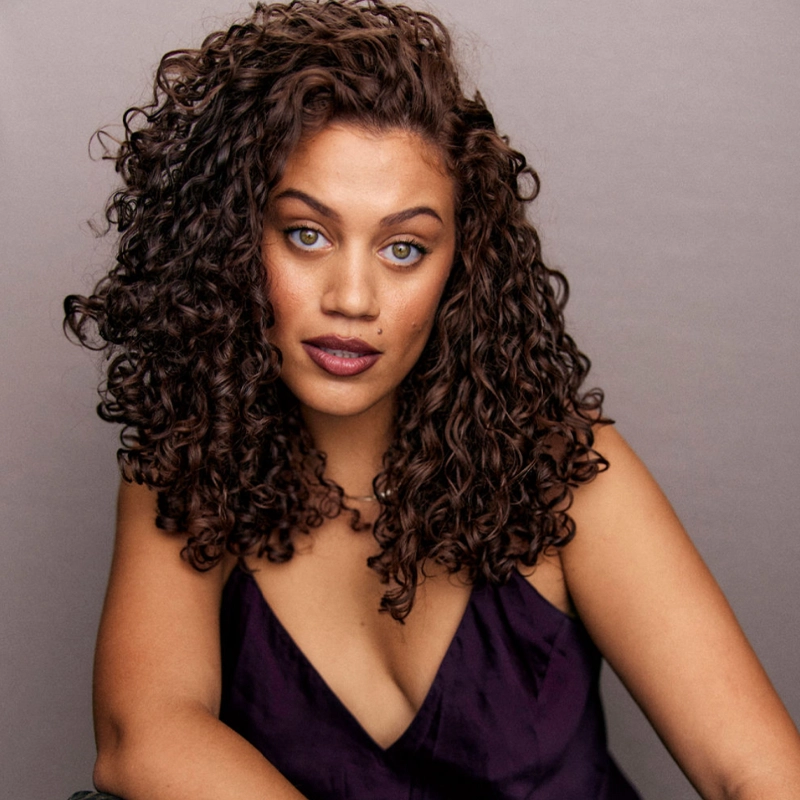 A woman with light brown skin and striking green eyes has shoulder-length, voluminous curly hair. She wears a deep purple, sleeveless top with a V-neck and poses confidently against a plain, light gray background, gazing directly at the camera.