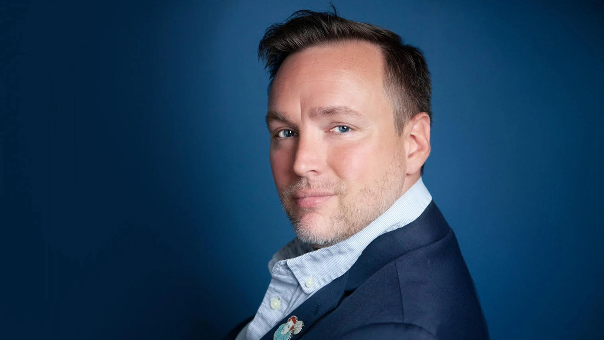 Robbie Rozelle - Show Image A confident man with short brown hair and light stubble, wearing a navy blue blazer over a light blue collared shirt, looks at the camera against a smooth, gradient blue background. With a slight smile, he channels cabaret flair in true Robbie Rozelle style.