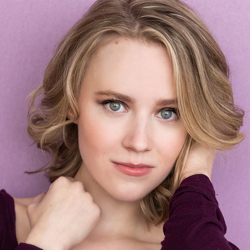 A young person with light skin and wavy blonde hair poses in front of a lavender background. They have blue-green eyes, subtle makeup, and wear a burgundy top. One hand touches their hair as they look directly at the camera with a soft expression.
