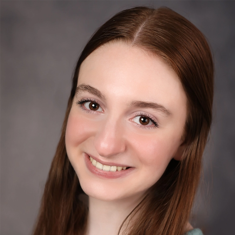 A young woman with long straight brown hair smiles at the camera. She has fair skin, brown eyes, and is wearing minimal makeup. The background is a soft, out-of-focus grey, drawing attention to her friendly expression.
