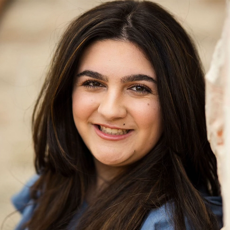 Samantha Kotok, a young woman with long, dark brown hair, smiles warmly at the camera. She has fair skin, dark eyebrows, and is wearing a blue top. The background is softly blurred, highlighting her cheerful expression.