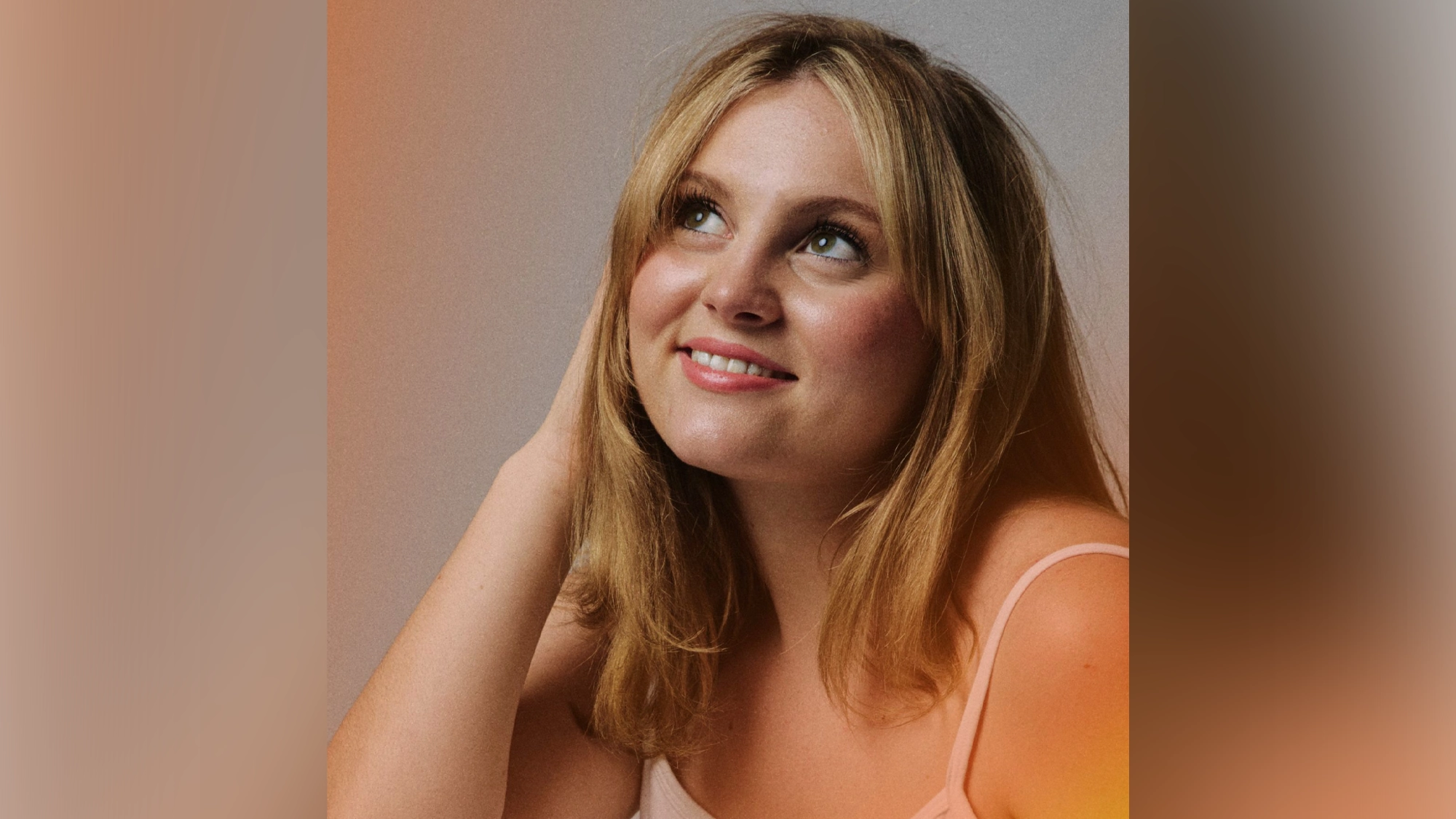 A woman with shoulder-length blonde hair smiles softly and looks up to the right. Samantha Stratton wears a light-colored tank top and touches her hair with one hand. The gentle lighting highlights her relaxed expression in this All Before 20 portrait.