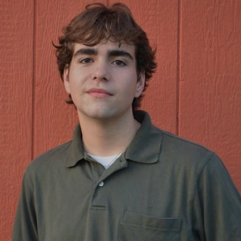 A young man with wavy brown hair and fair skin stands in front of an orange wooden wall. Lukas Schoellkopf wears a green collared shirt over a white undershirt and looks calmly at the camera with a neutral expression.