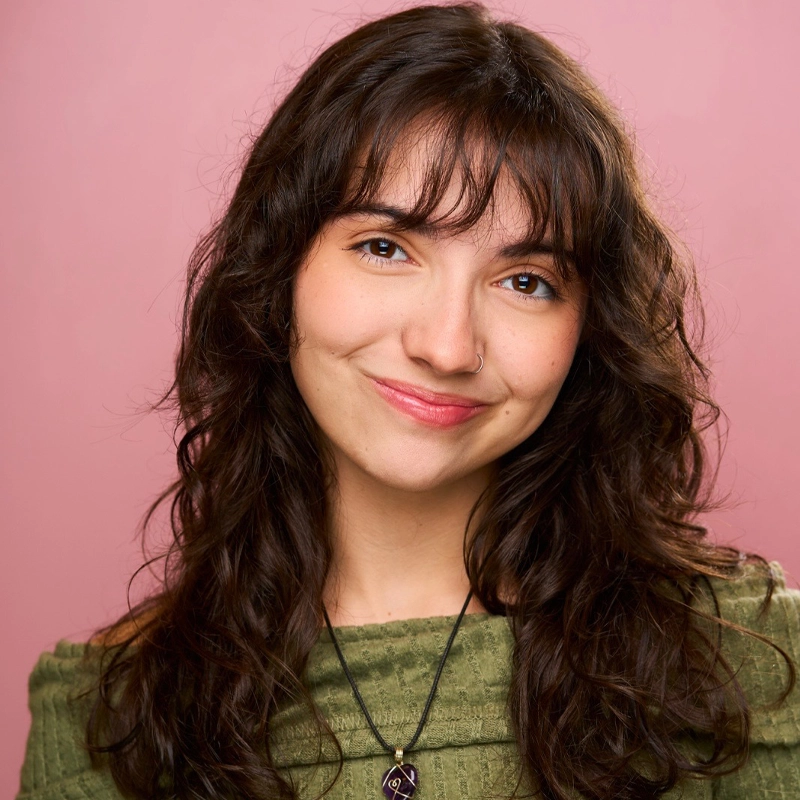 A young woman with wavy dark brown hair and bangs smiles softly in front of a pink background. She wears a green textured top, a nose ring, and a necklace with a purple crystal pendant. Her expression is warm and friendly.