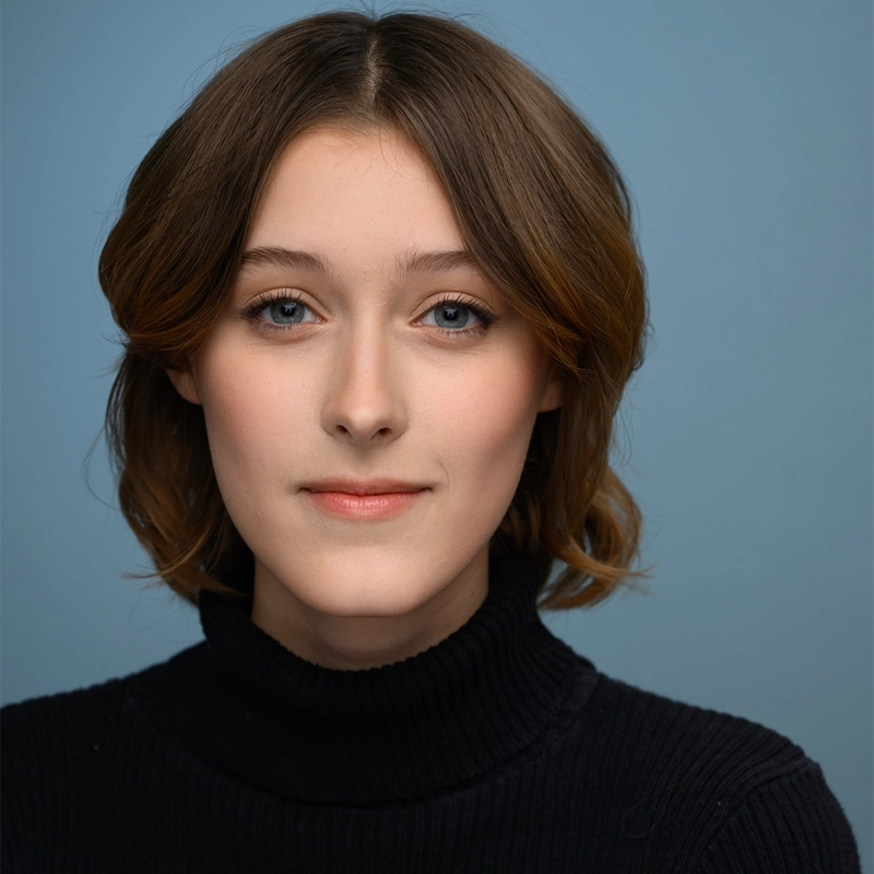 A young woman with fair skin, short brown hair, and blue eyes wears a black turtleneck sweater. Riley Shroyer is facing forward, smiling softly, and posing against a plain light blue background with even lighting.