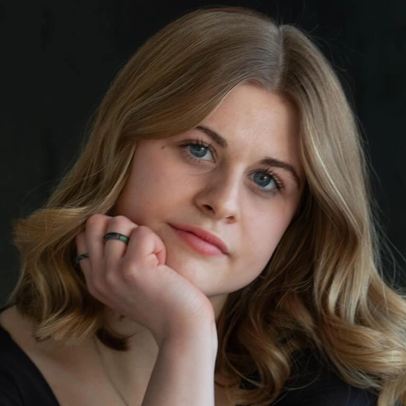 A young woman with wavy, light brown hair rests her chin on her hand. She looks directly at the camera with a neutral expression. She wears a black top and a ring on her right index finger. The background is dark and out of focus.