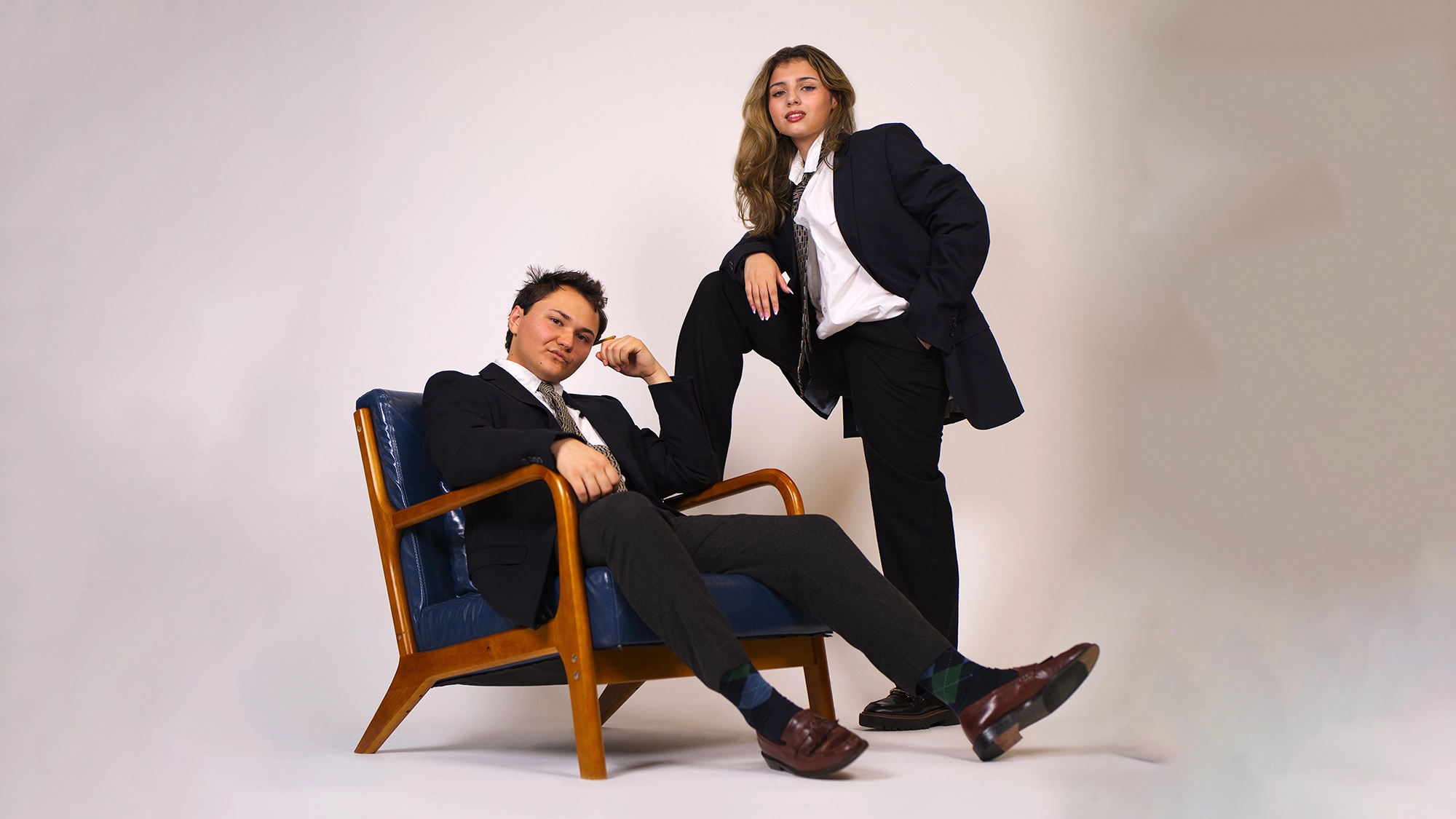 Sparr and Gallo site image 26 Meg Gallo and Grant Sparr pose confidently in a studio with a plain background. One sits relaxed in a vintage blue chair while the other stands beside them, both dressed sharply—ready to ring in the New Year with style.
