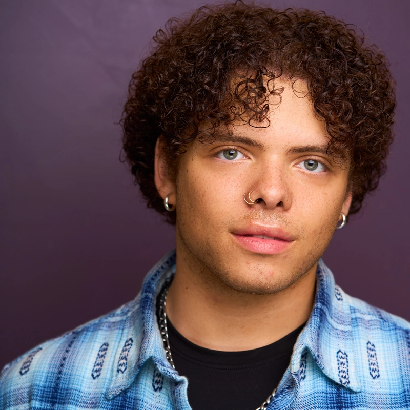 A young person with light brown skin, blue eyes, and short curly brown hair stands against a purple background. They wear a blue plaid shirt over a black top, silver chain necklace, and have a nose ring and hoop earrings, looking confidently at the camera.