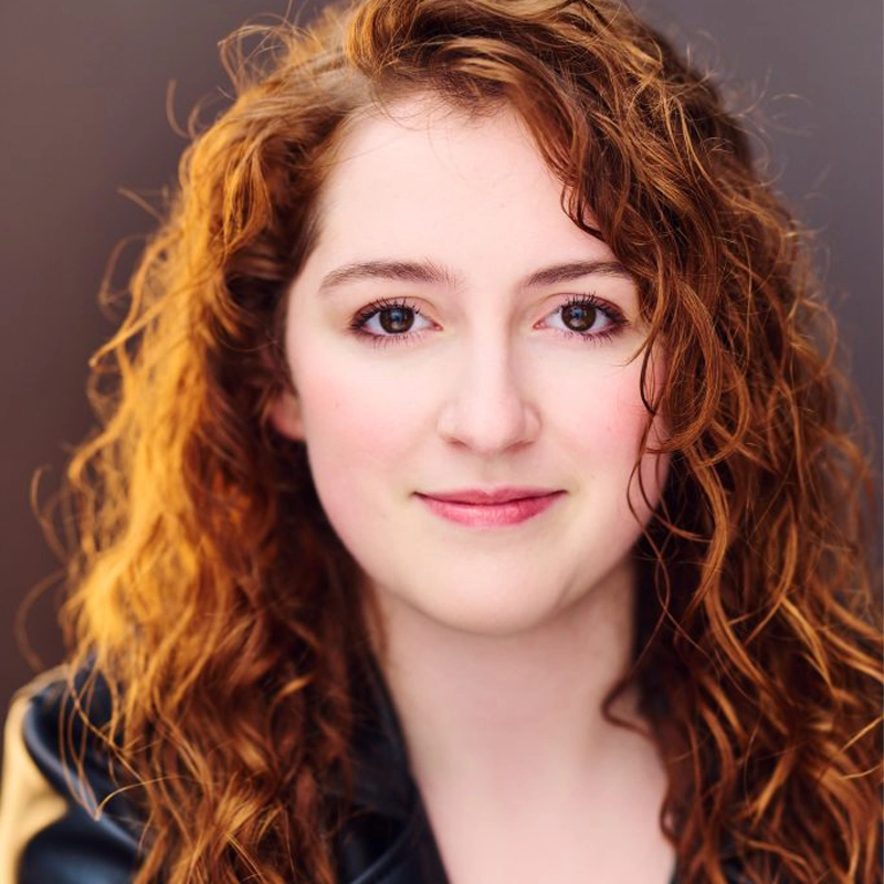 A young woman with fair skin and long, curly red hair, Skye Stauffer, looks directly at the camera, softly smiling. She has brown eyes and is wearing a black jacket. The blurred, neutral background draws attention to her face.