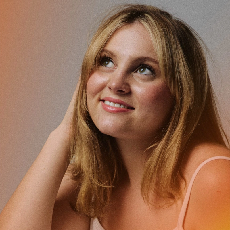Samantha Stratton, a young woman with blonde hair and fair skin, smiles softly as she looks up and to the side. She wears a light pink sleeveless top, touching her hair, with a warm orange light effect at the edges of the neutral background.