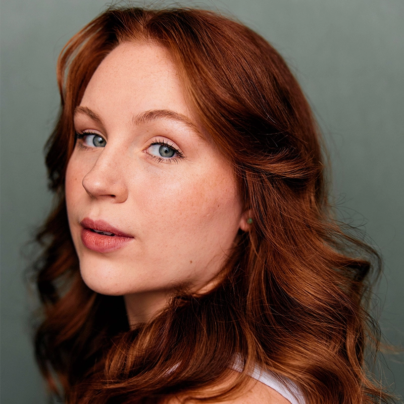 A young woman with fair skin, wavy red hair, and blue eyes is gazing slightly over her shoulder at the camera. She has a calm, neutral expression and wears a light-colored top. The background is a plain, muted teal color.