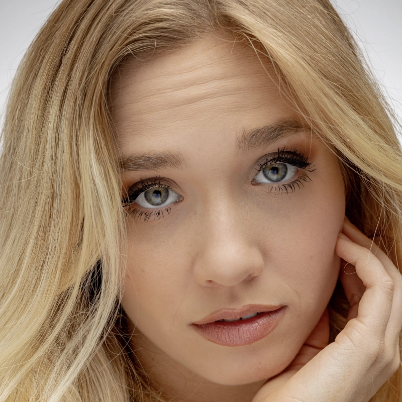 Chloe Vallot, a young woman with long blonde hair and blue eyes, looks directly at the camera. She gently rests her face on her hand, wearing subtle makeup. The plain, light background keeps the focus on Chloe's serene expression.