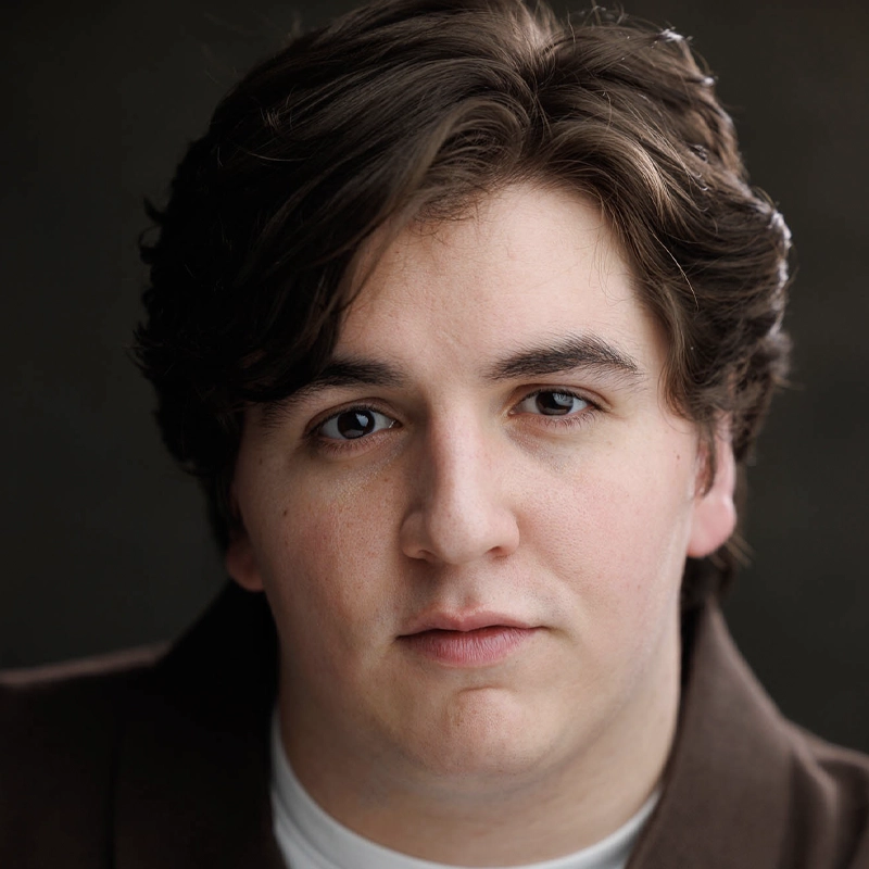 A young man with fair skin and medium-length, wavy brown hair looks directly at the camera. He has brown eyes and a neutral expression. He wears a brown jacket over a white shirt. The background is softly blurred and dark.