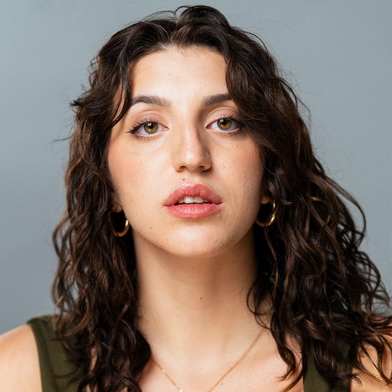 Vita Augugliaro, with wavy dark brown hair and hazel eyes, looks directly at the camera against a plain gray background. She wears a sleeveless olive top, gold hoop earrings, and a delicate necklace, her expression calm and neutral.