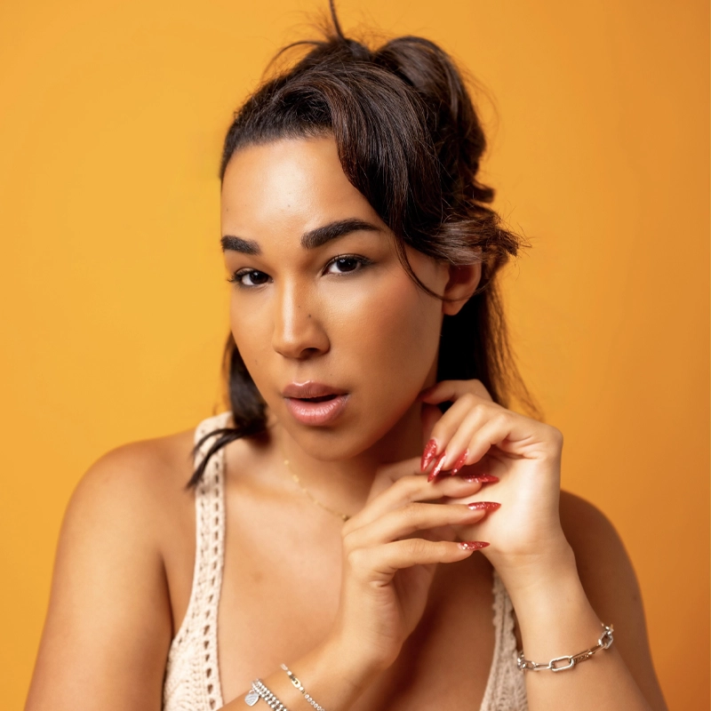 A woman with long dark hair styled half-up poses against an orange background. She wears a cream crochet top, silver bracelets, and has long red nails. She gazes at the camera with her hands gently touching her chin and collarbone.