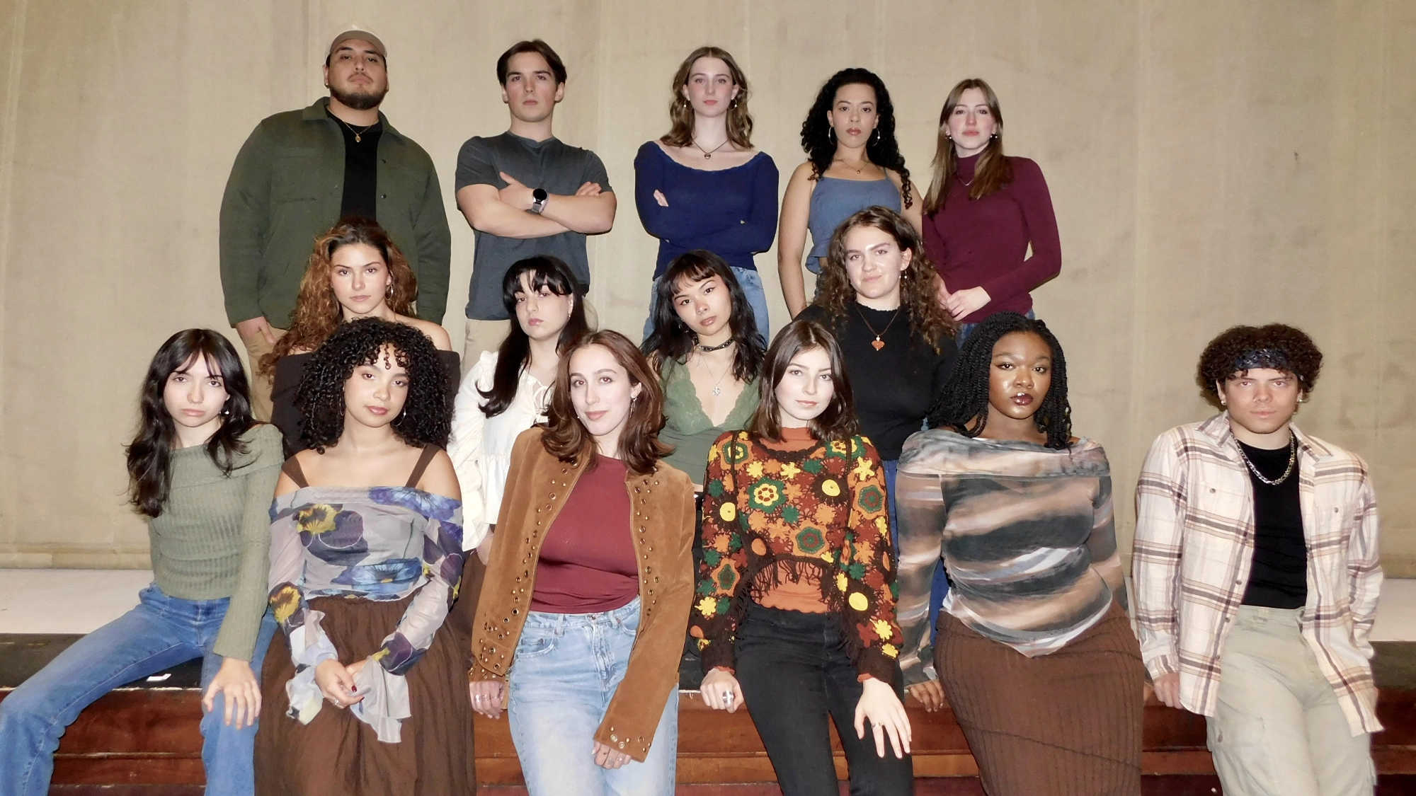 Fifteen young adults from the BFA Musical Theatre Class of 2026 at Western Connecticut State University pose indoors in two rows against a plain beige wall, dressed in stylish fall outfits and gazing at the camera with neutral expressions.