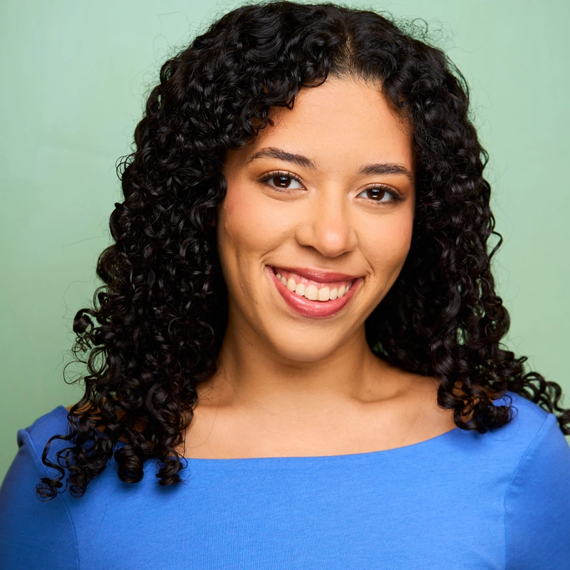 Savannah Wright, a woman with medium skin tone and long, curly dark hair, smiles at the camera. She is wearing a bright blue top and has natural makeup. The background is a solid, light green color.